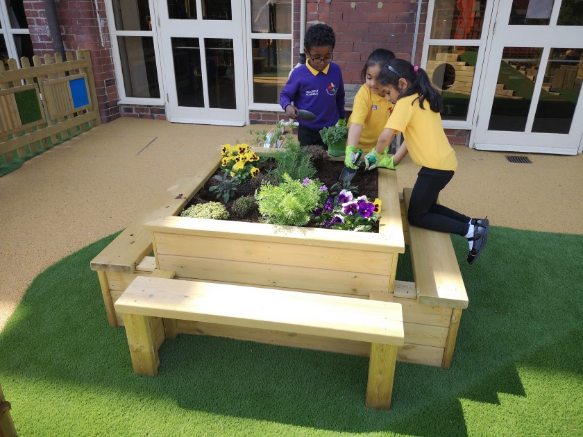 3 children are planting flowers and placing soil in a wooden planter. The planter has 4 wooden benches attached to it, allowing for the children to kneel on it to dig the soil. 2 children are holding garden trowels