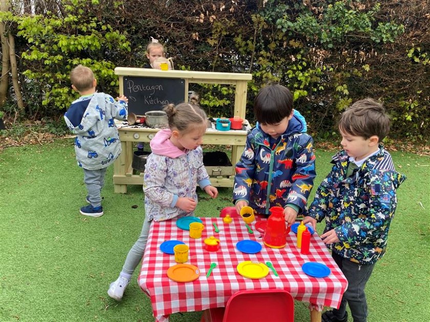 3 children are stood at a dinner table with toy plates and cutlery, whilst one child is operating the mud kitchen and creating some imaginary food. All 4 children are taking part in EYFS role play activities.