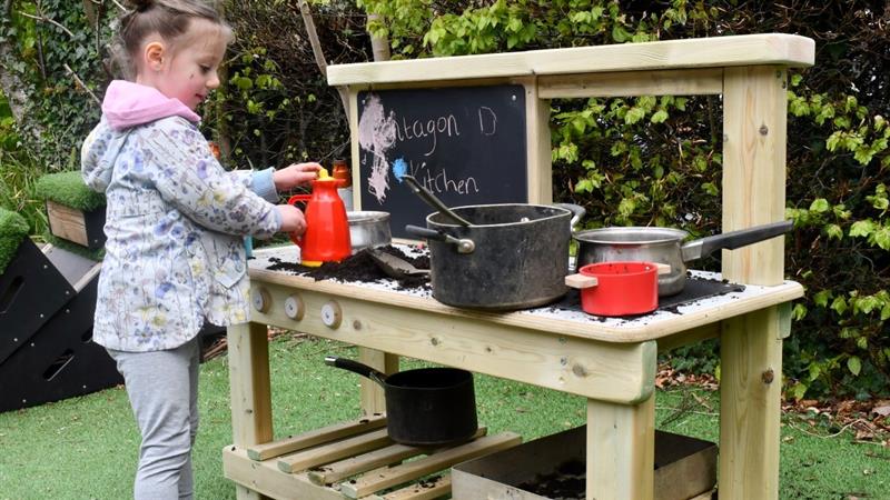 a little girl stands at the essentials mud kitchen and mixes things with the toys 