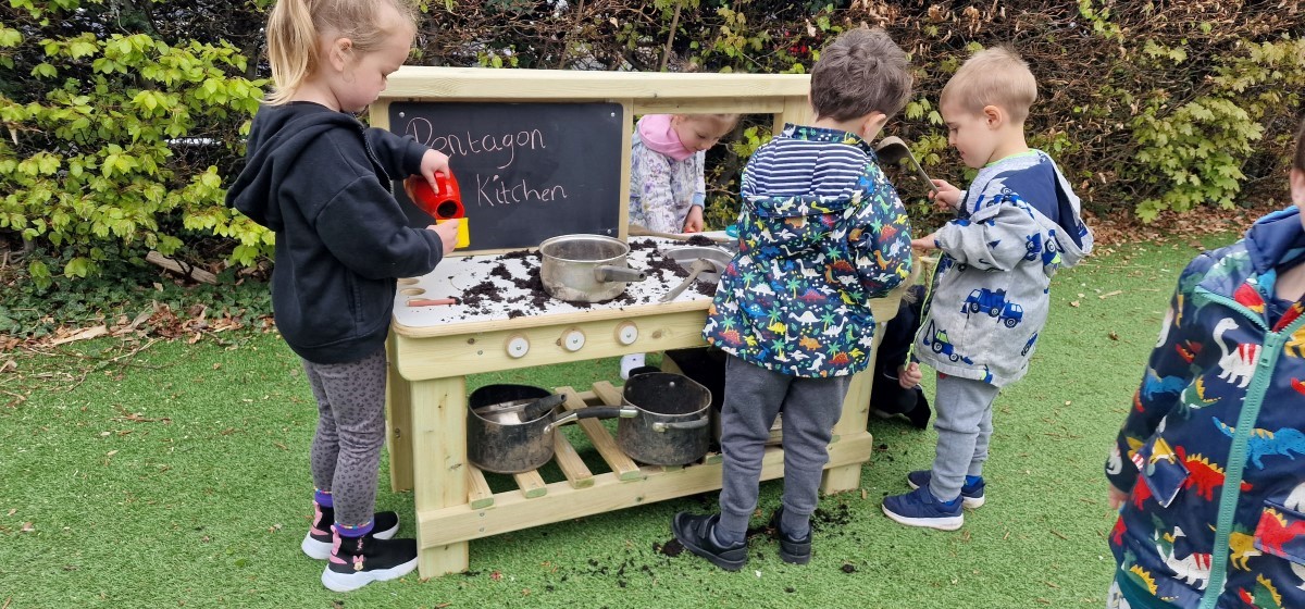 A group of 4 children surrounding the outdoors mud kitchen, designed by Pentagon Play. The four children are using pretend play to create dishes with the mud and on the chalkboard, built onto the mud kitchen, someone has wrote "Pentagon Kitchen"