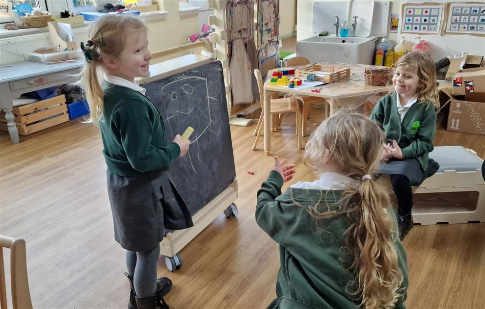 A little girl is stood beside a movable chalkboard and is talking to two of her classmates. The two other girls are watching the girl near the chalkboard and are smiling.