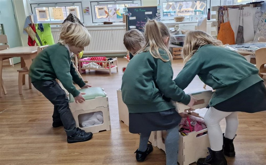 4 Children are opening the stack and sit stools and are retrieving role play outfits from them. They are getting ready for pretend play as a wooden crib can be seen in the background. 