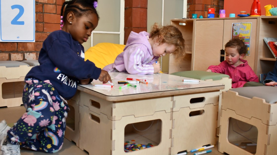 A group of 3 children are sat at a whiteboard table and are drawing a variety of pictures with each other.