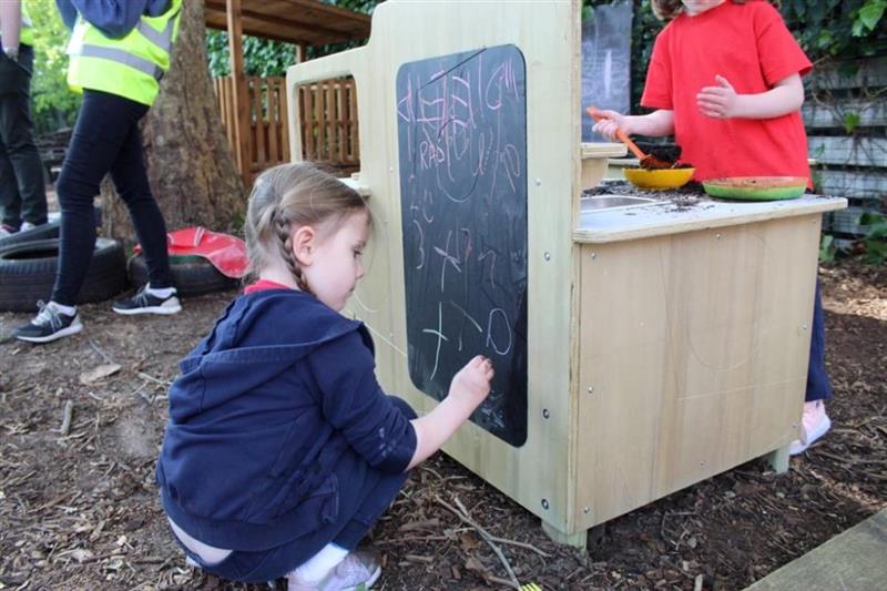 a child kneels on the floor round the back of the mud kitchen and draws on the chalkboard