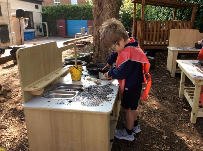 a little boy stands at the mud kitchen and mixez and makes with different baking props