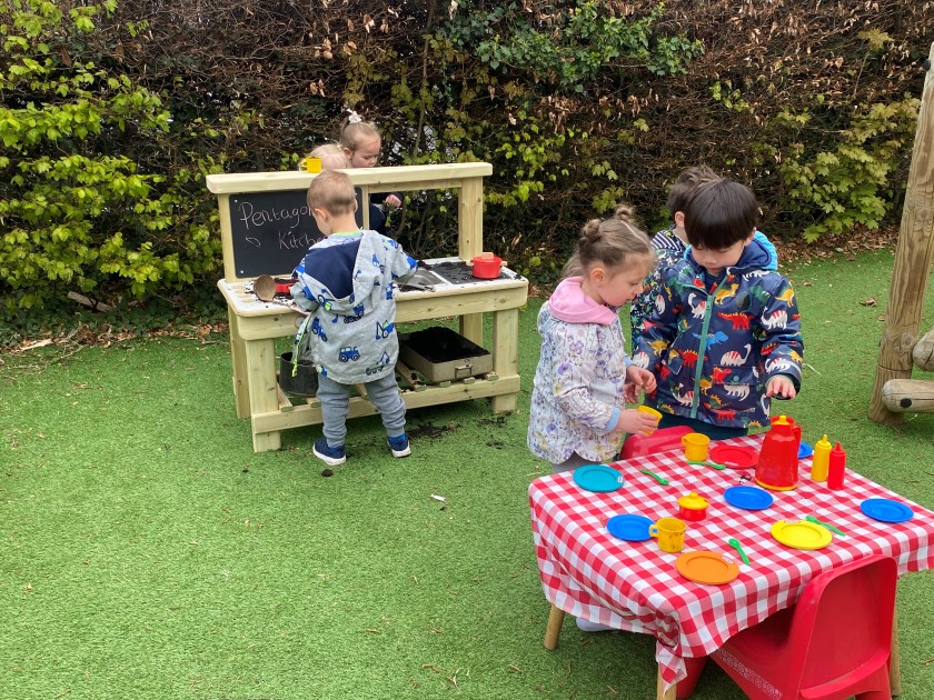 Tree children are playing with a Mud Kitchen whilst another three are setting up a dinner table. One child is watching this all happen.