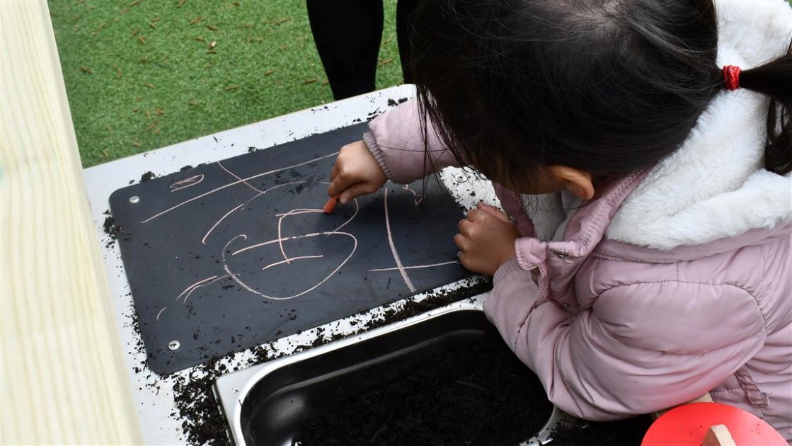 A child drawing on a chalkboard, which is attached to the Mud Kitchen product (designed by Pentagon Play). The child is scribbling pink chalk on the board