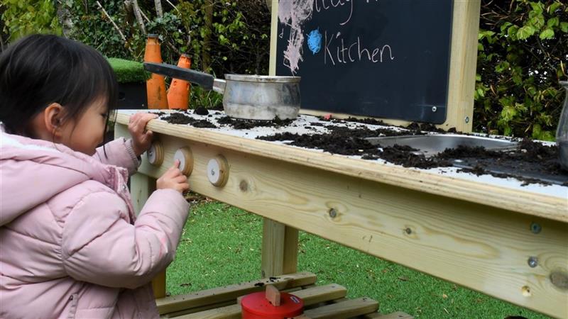 a little girl kneels in front of the mud kitchen and twiddles with the dials