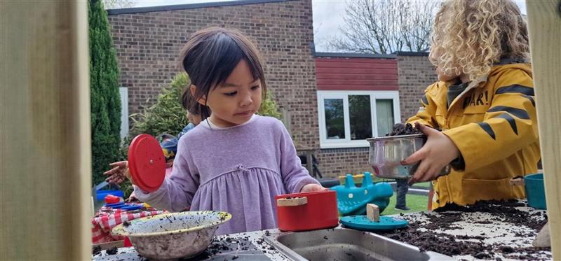 a little girl stands and looks into a pot on the mud kitchen surface