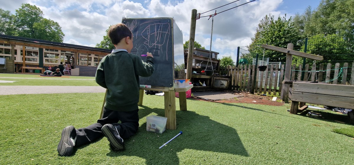 A boy is drawing on the Group Art Easel, supplied by Pentagon Play. The boy is sketching his own design with pink chalk as the sun is shining on him.