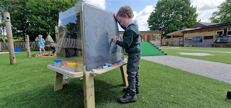 a child stands and leans on the group art easel as he draws on the chalkboard panel of the group art easel