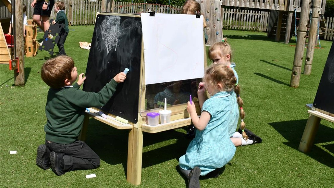a group of 4 children are surrounding the group art easel from our essentials range. The children are using chalk to draw on the chalkboard whilst one child has a piece of paper attached to the chalkboard and is painting on it.