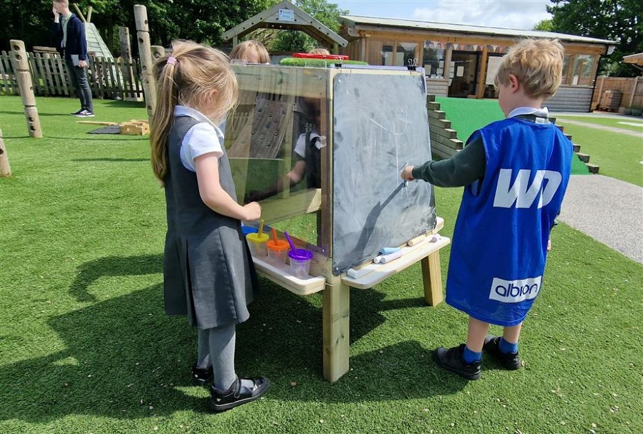 Three children are engaging with the group art easel. A boy is wearing a blue jacket as he draws on the chalkboard, whilst the little girl to his left is looking at all the paints she can use. The other child is at the back of the easel, drawing on a chalkboard. The group art easel is set up on an artificial grass top.