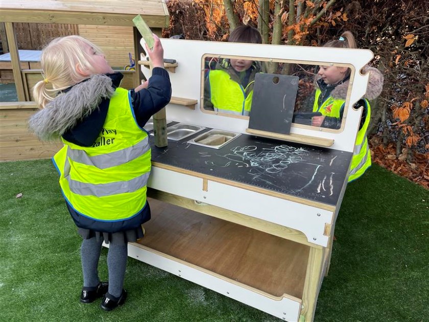 3 children are stood around a wooden work table and are wearing hi-vis jackets. One little girl is holding a wooden block in the air and is looking at it's size. The other two children are stood on the other side and are measuring other objects,