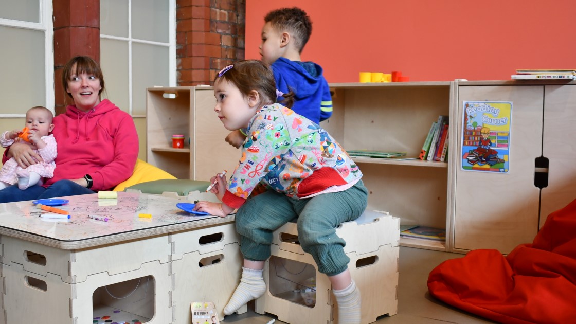 2 children are drawing on a whiteboard whilst a parent sits in a seat and holds a baby.