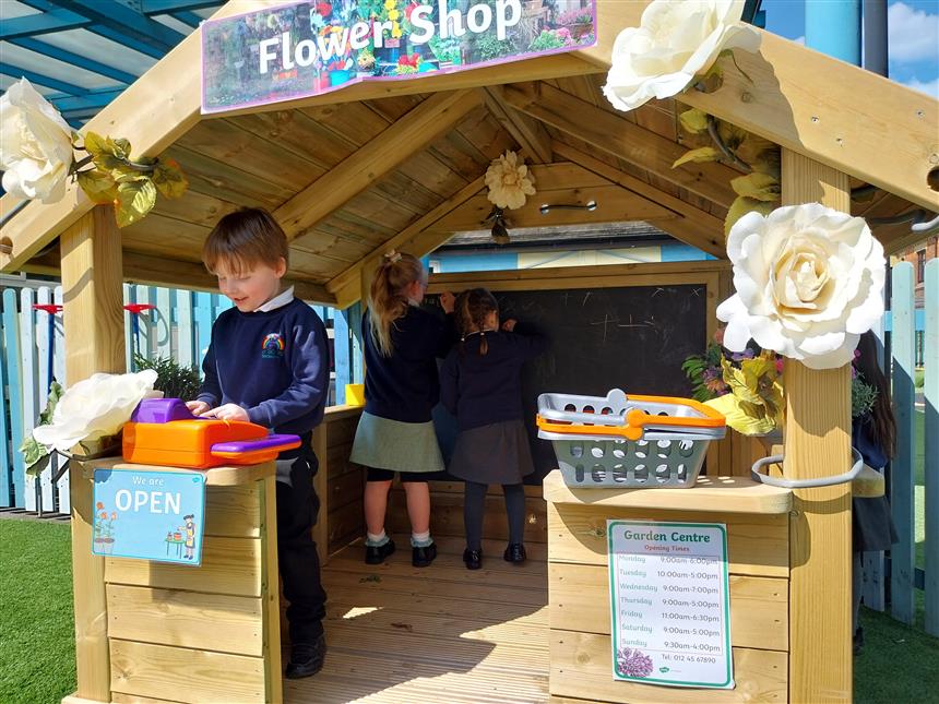 Three children are in a wooden playhouse which has a banner at the top saying "flower shop". One child is stood at the front, operating a toy till, whilst two girls are at the back of the playhouse, drawing something on the big chalkboard.