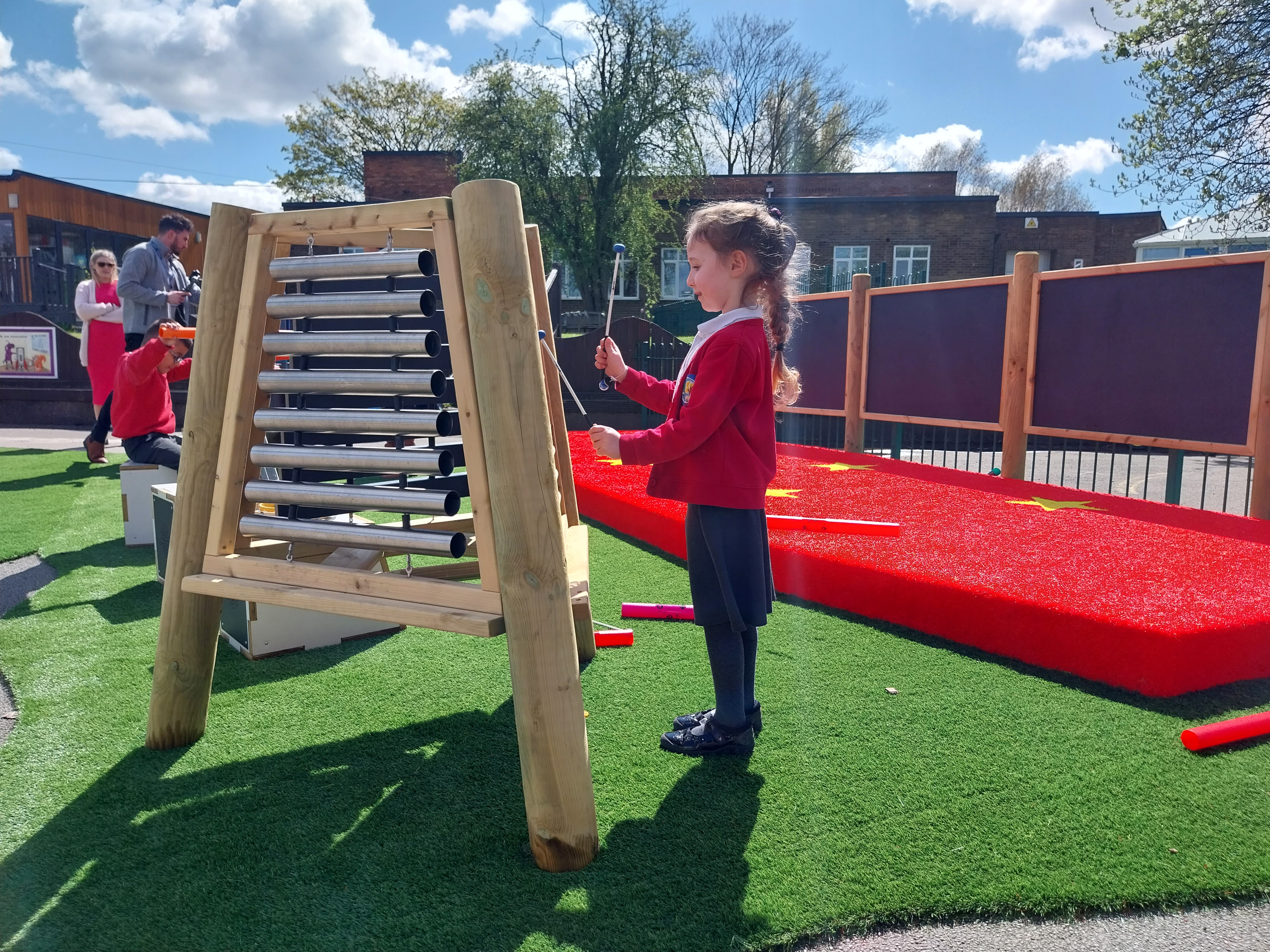 A little girl is playing a freestanding marimba on an artificial grass surface. An outdoor stage with chalkboards can be seen behind her and a group of children playing in the distance.