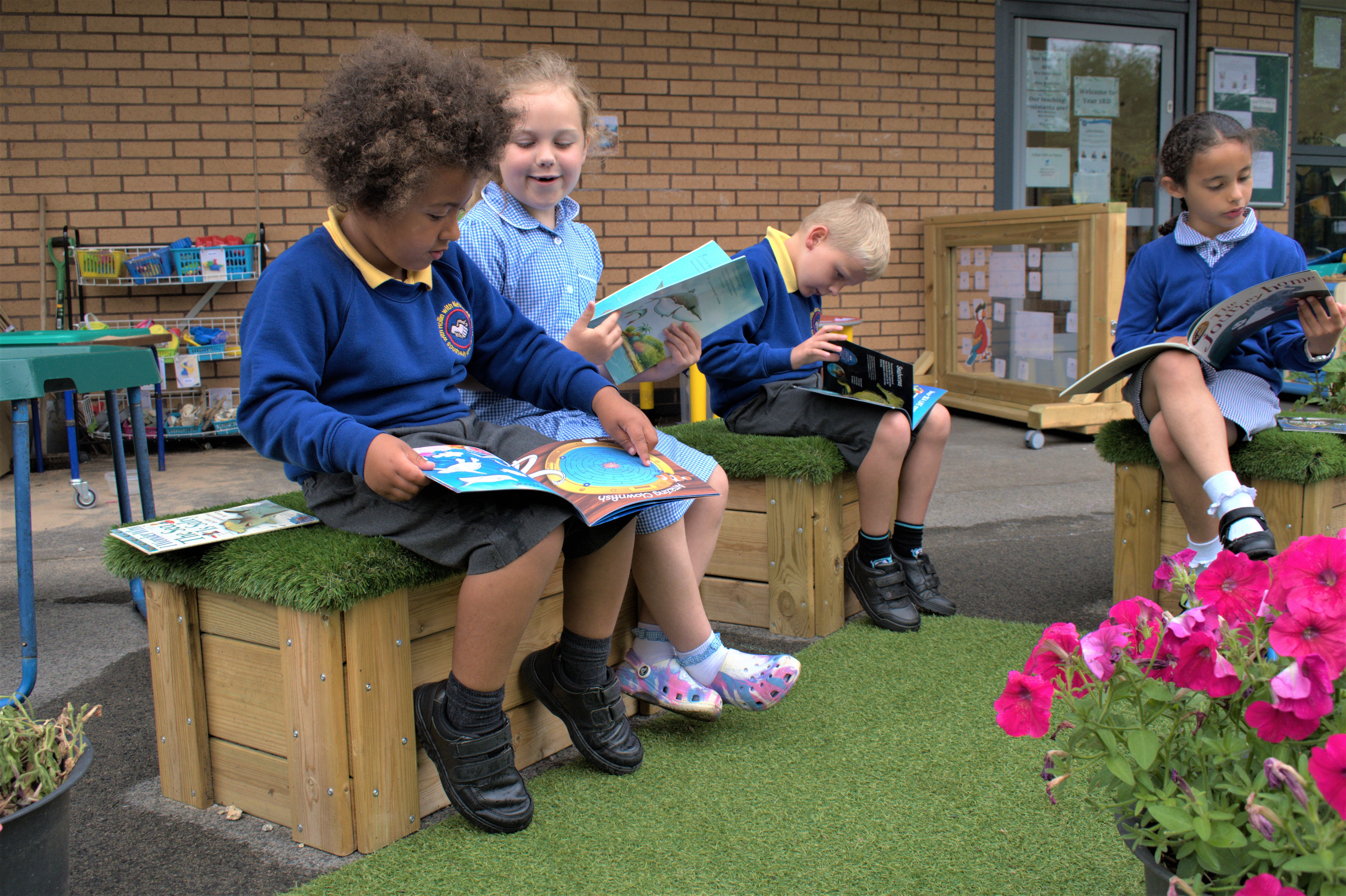 A group of four children are sat on artificial grass topped seats outside as they are reading books.