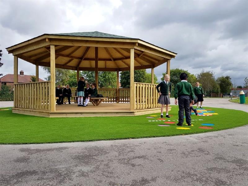 children play on the playground markings and children sit and work together under the shelter of the wooden gazebo