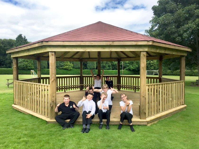 A group of kids are sat on the wooden flooring of a huge gazebo. The main structure of the gazebo is made out of wood, with a red roof made from shingles. The children are sat in the doorway and are looking at the camera, smiling.