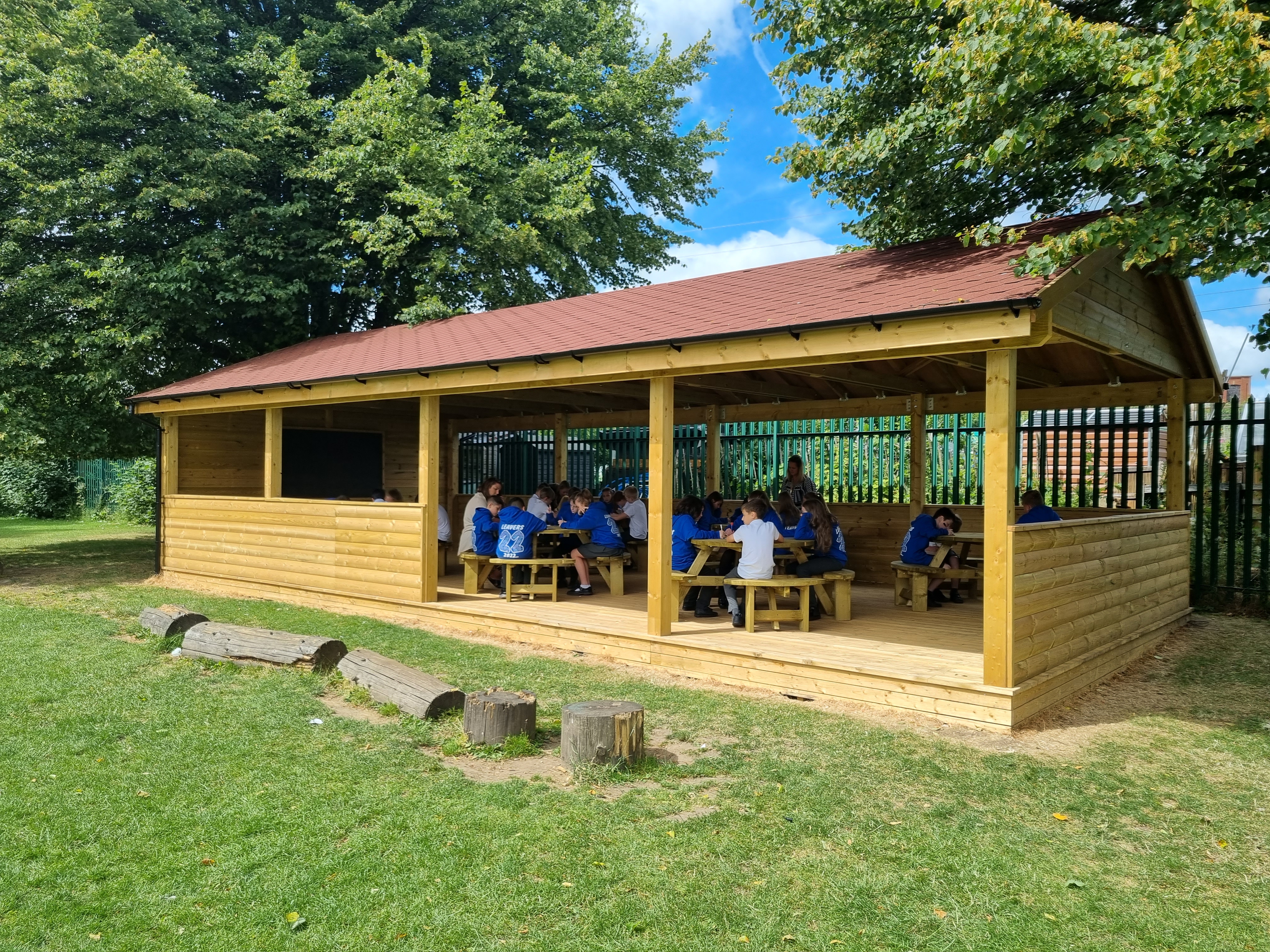 A wooden gable-end canopy with a class of children inside, studying on circular tables.