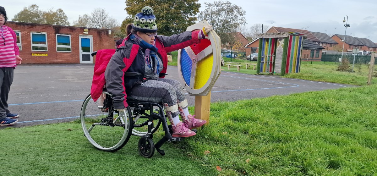 A girl in a wheelchair is playing with a sensory spinner, which is attached to a wooden pillar that has been buried into the ground.