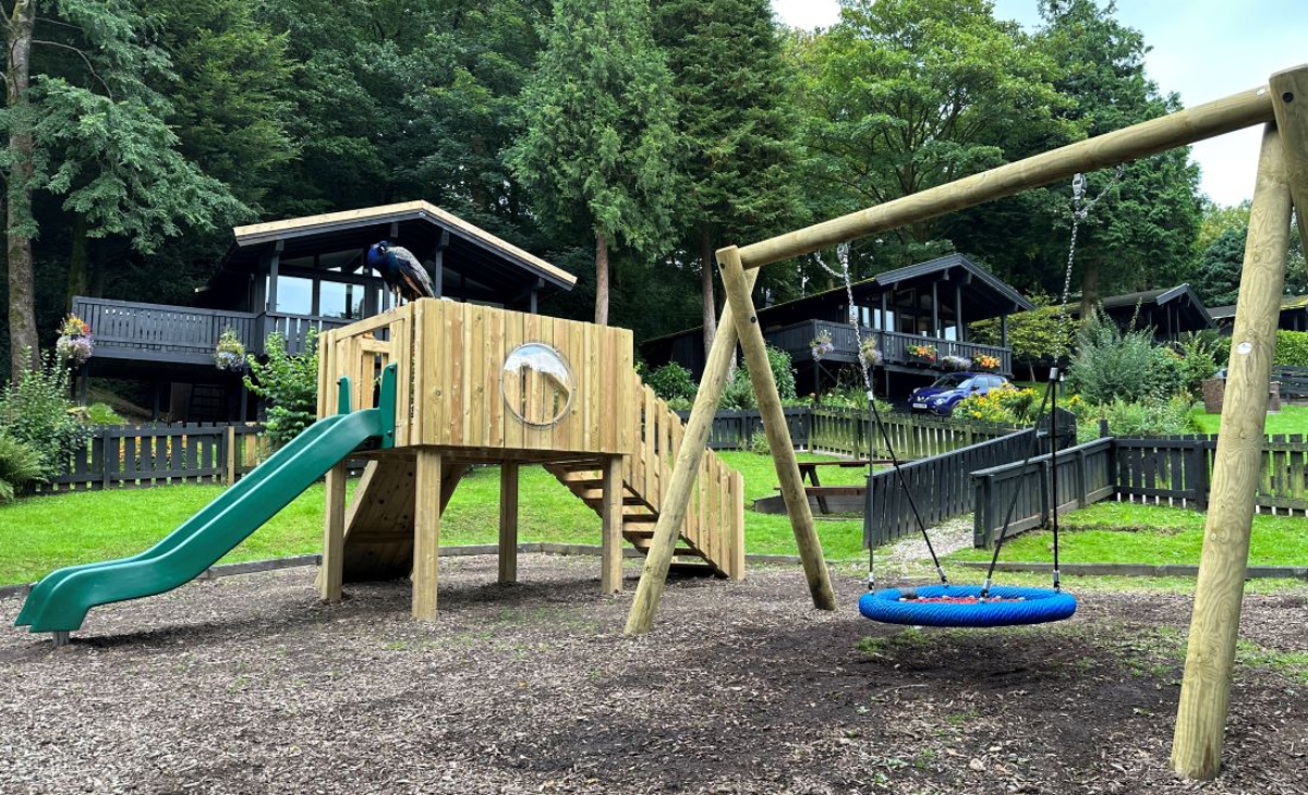 A play tower with a set of stairs, a climbing wall and a green slide with a basket swing next to it. The flooring is wooden bark. A dense forest is surrounding the park, with a few cabins looking over it.