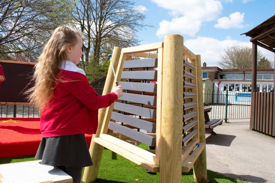 A little girl is playing with a piece of freestanding musical equipment on the playground.