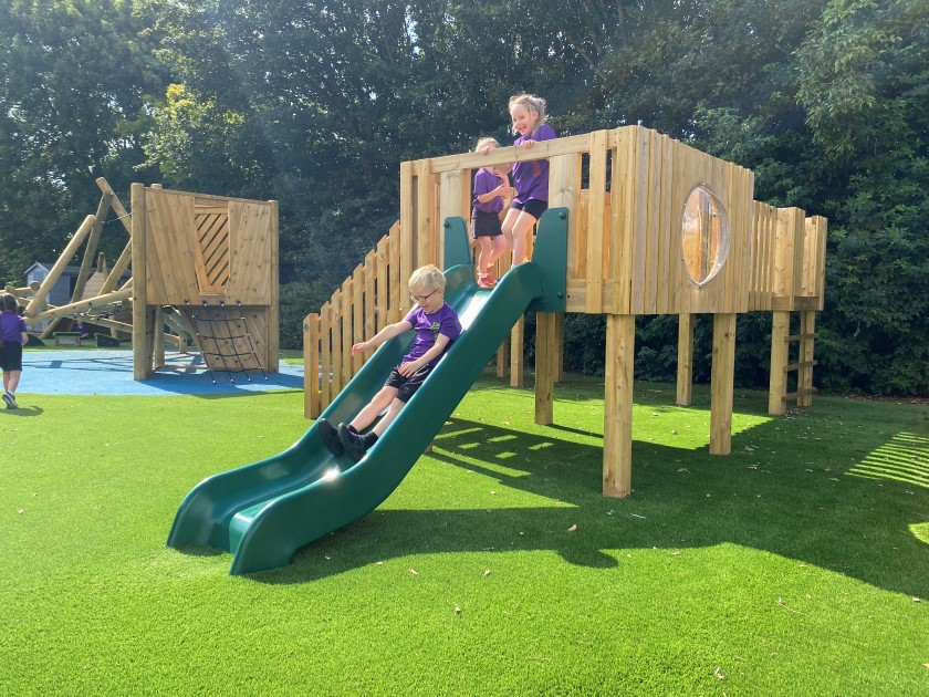 A Play Tower that is made of wood and has a plastic slide and window. Three children are playing with the equipment and are staying active. The equipment has been installed on artificial grass and is next to a climbing wall.