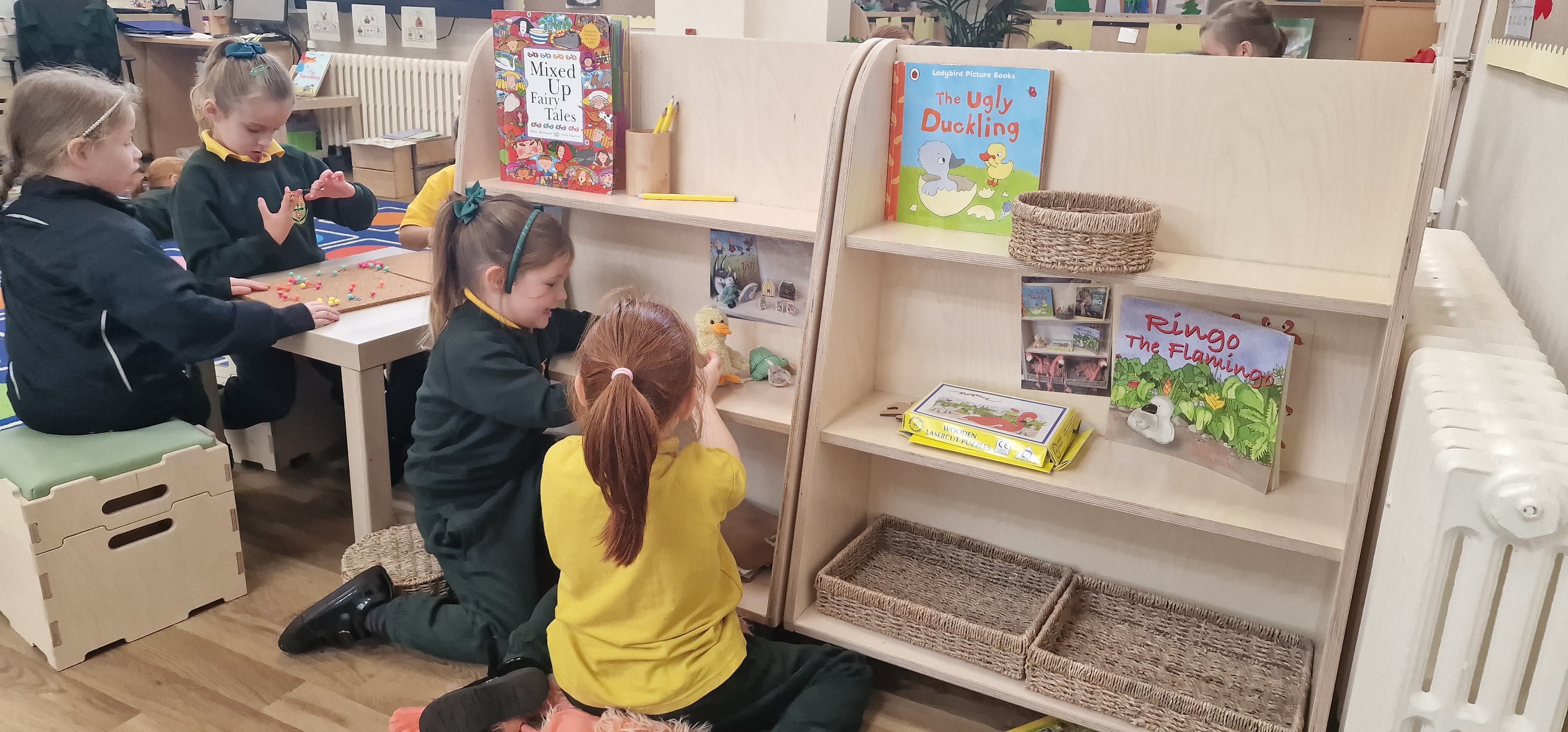 2 children are looking through the angled storage on wheels, created by Pentagon Play. In this indoor classroom, the storage unit is displaying books, teddies and pencils.