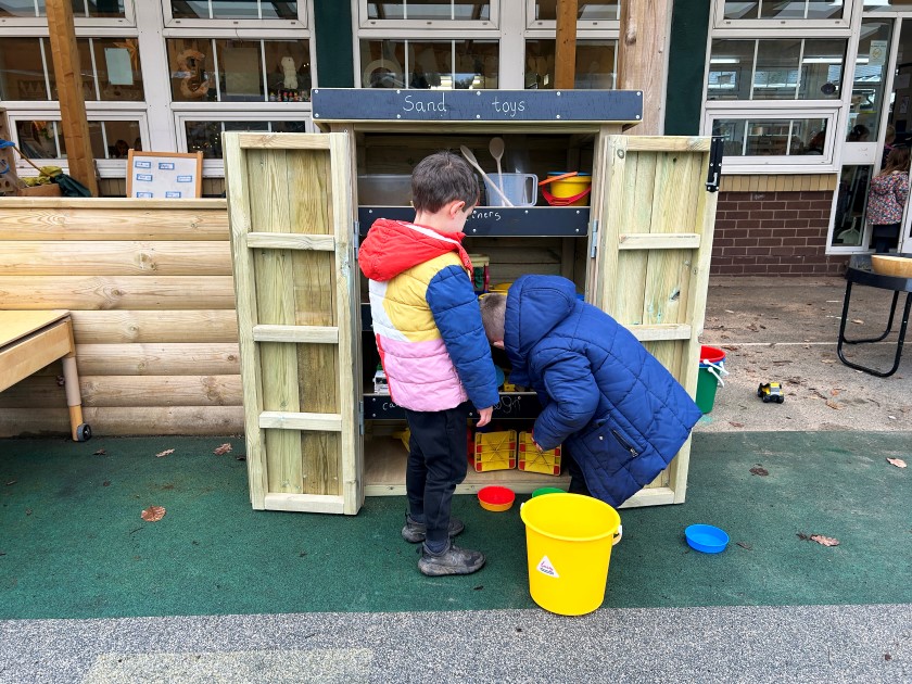 2 children are looking through one of Pentagon Play's Acorn Self-Select Stores. In this photo, the Acorn Self-Select store is being used to store sand toys and other creative play equipment. On the top of the Acorn storage unit, the words "Sand Toys" have been written on the chalk board.