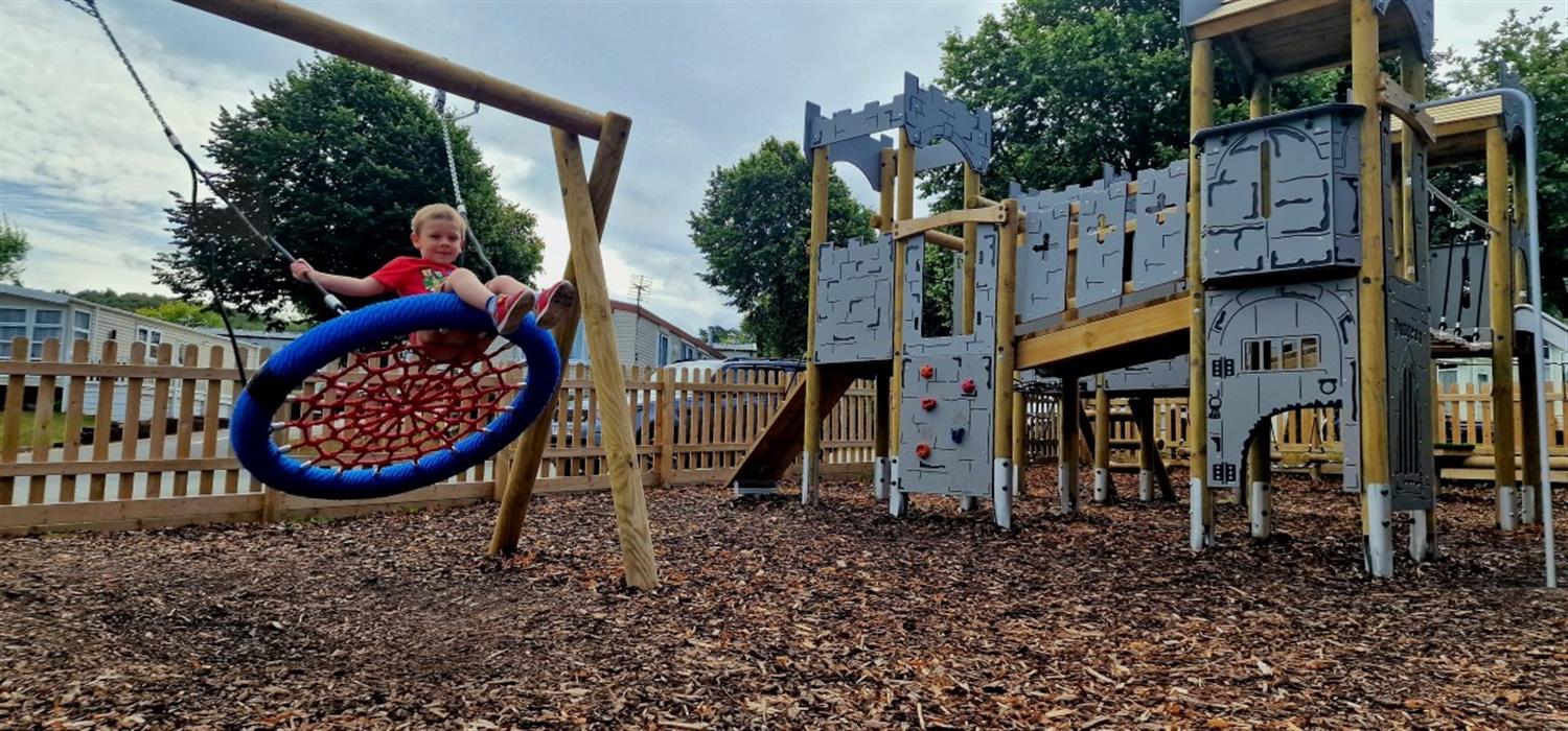 A photo showing a child on a swing, with a play frame in the background. The whole play area has been designed by Pentagon Play