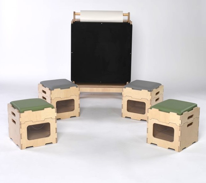 4 stack and sit blocks are placed in front of Pentagon Play's indoor art easel. 2 of the Stack and Sit blocks have a sage green seat cushion, where the other 2 have a grey cushion
