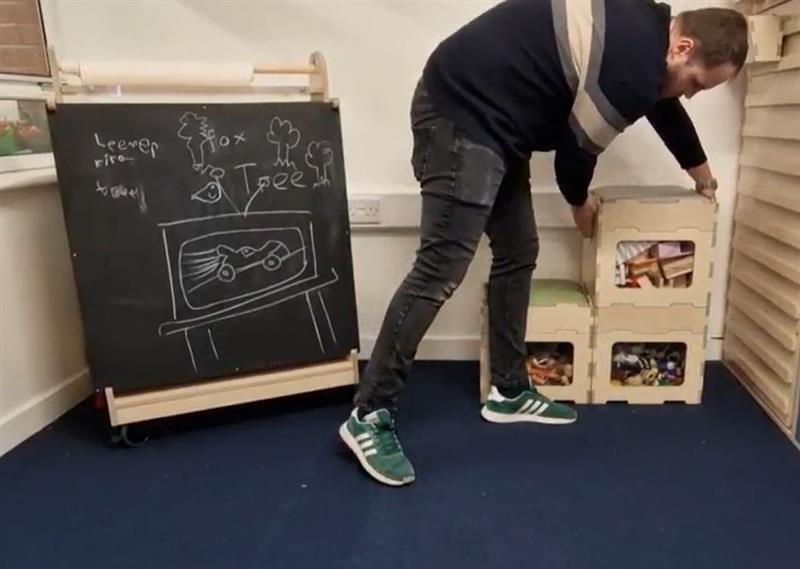 A teacher is stacking the "Stack and Sit blocks" on top of each other to create more room. To the left of him is an indoor art easel which has a lesson drawn on it. All this equipment was provided by Pentagon Play