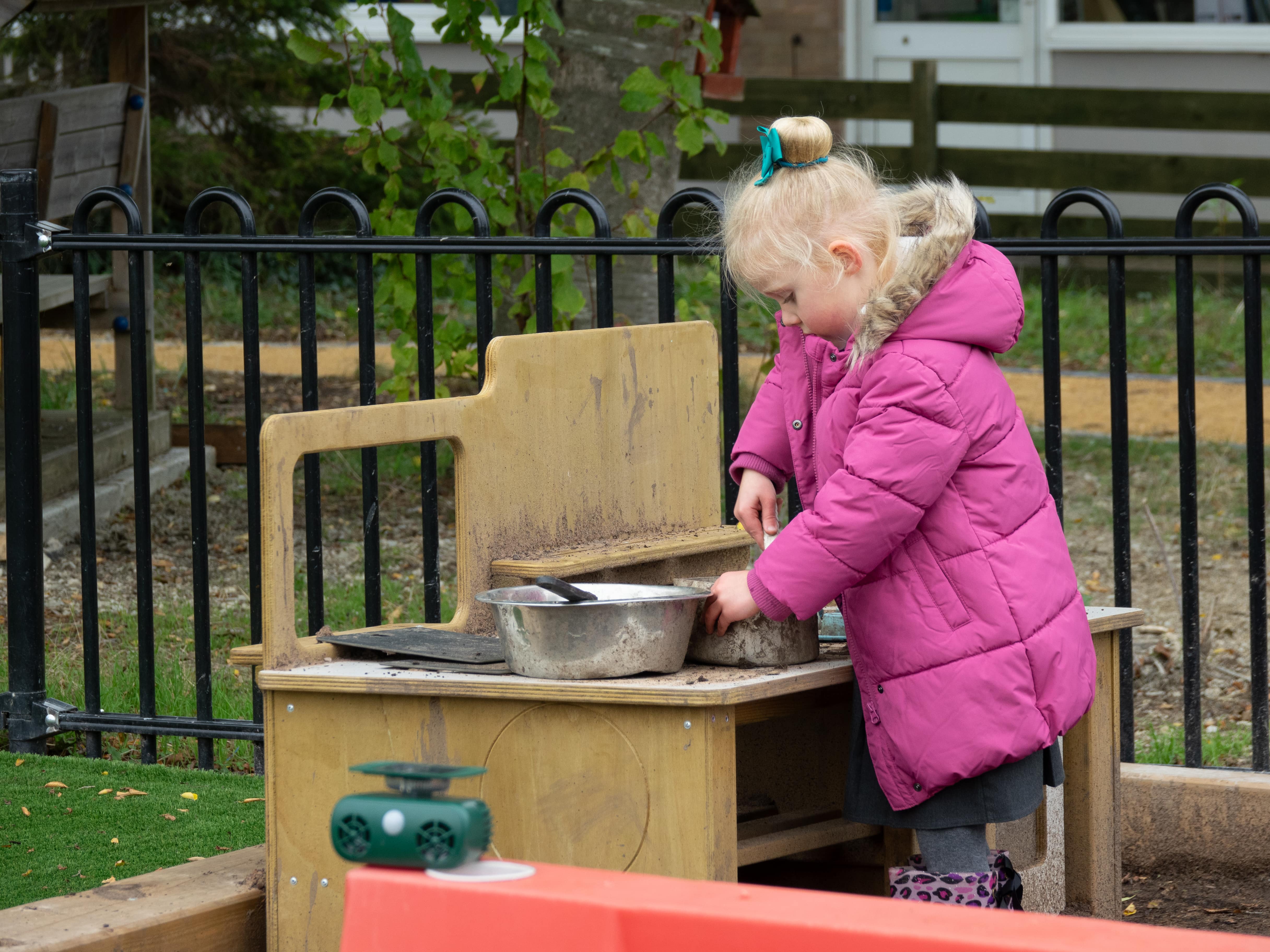A little girl wearing a pink coat is playing with the mud kitchen equipment, made by Pentagon Play. She is pouring mud into a pan and stirring it.