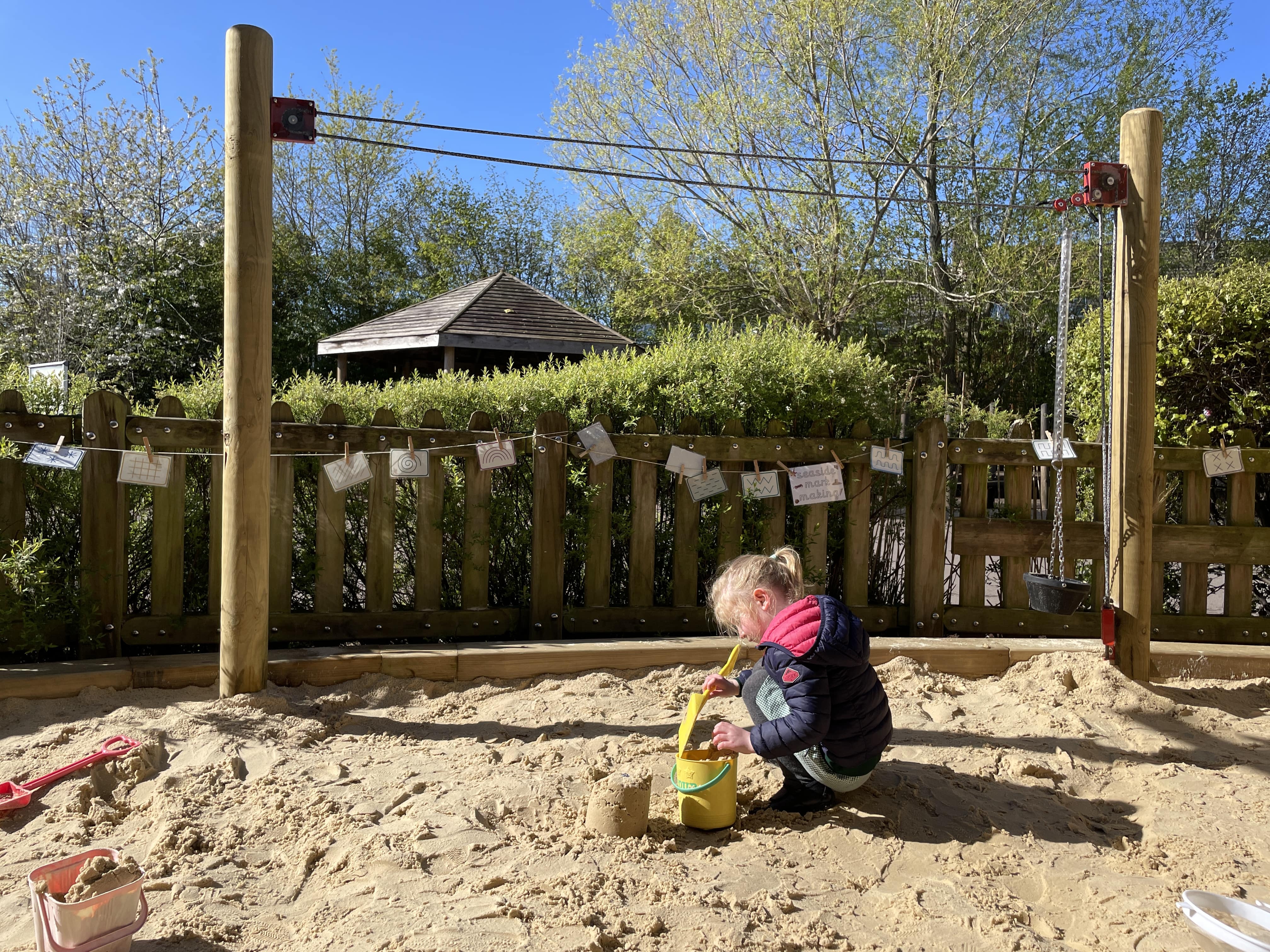 A little girl is in a sandpit with a plastic shovel and bucket, building sandcastles. Behind her is the rope and pulley play equipment, which is two wooden poles that are connected by rope and have a pulley attached, allowing buckets to be transported between the two.