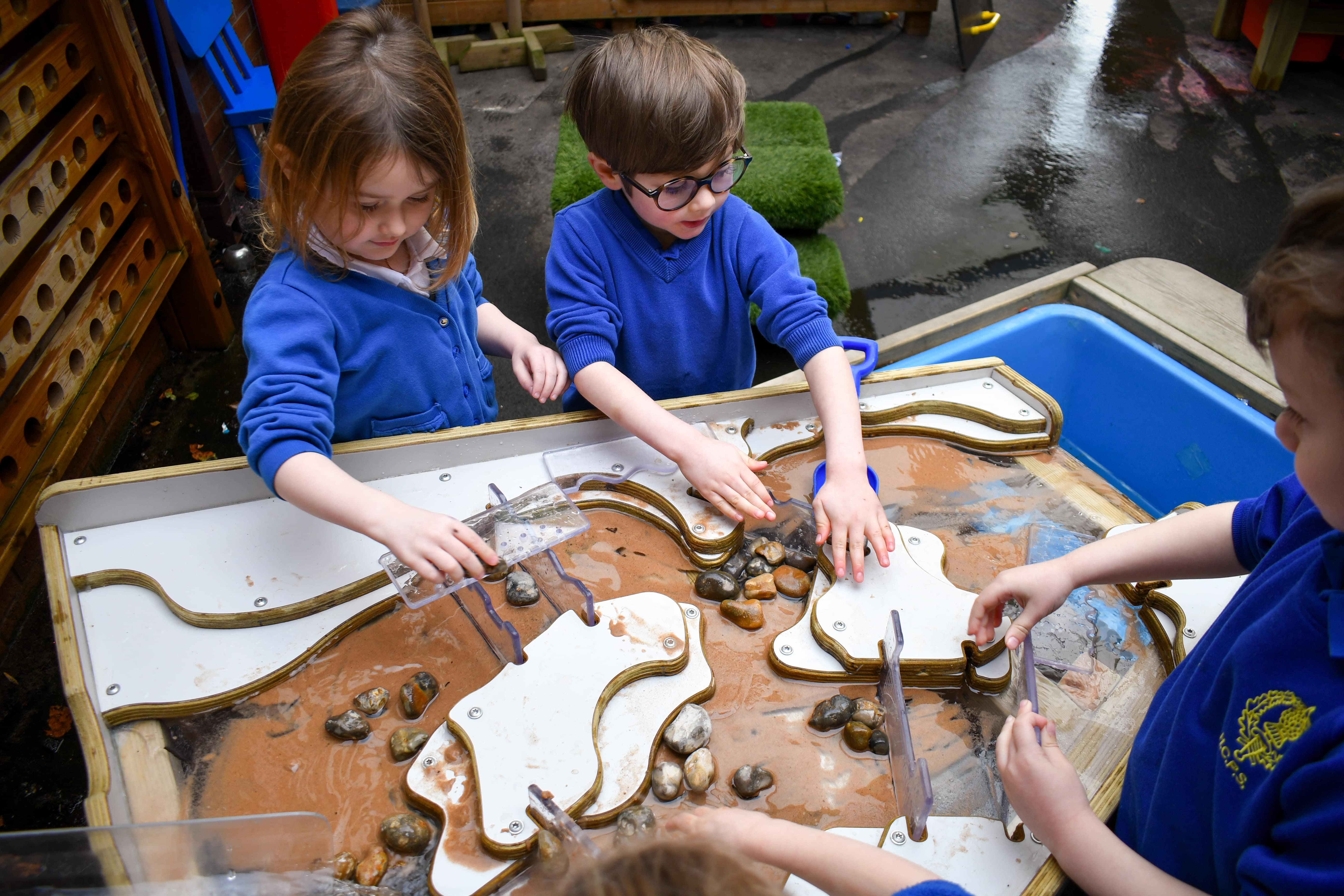 A group of four children are playing with Pentagon Play's Damming Station. Water is pouring down the damming stations table, as it hits pebbles and plastic water dams, eventually pouring into a bucket at the end.