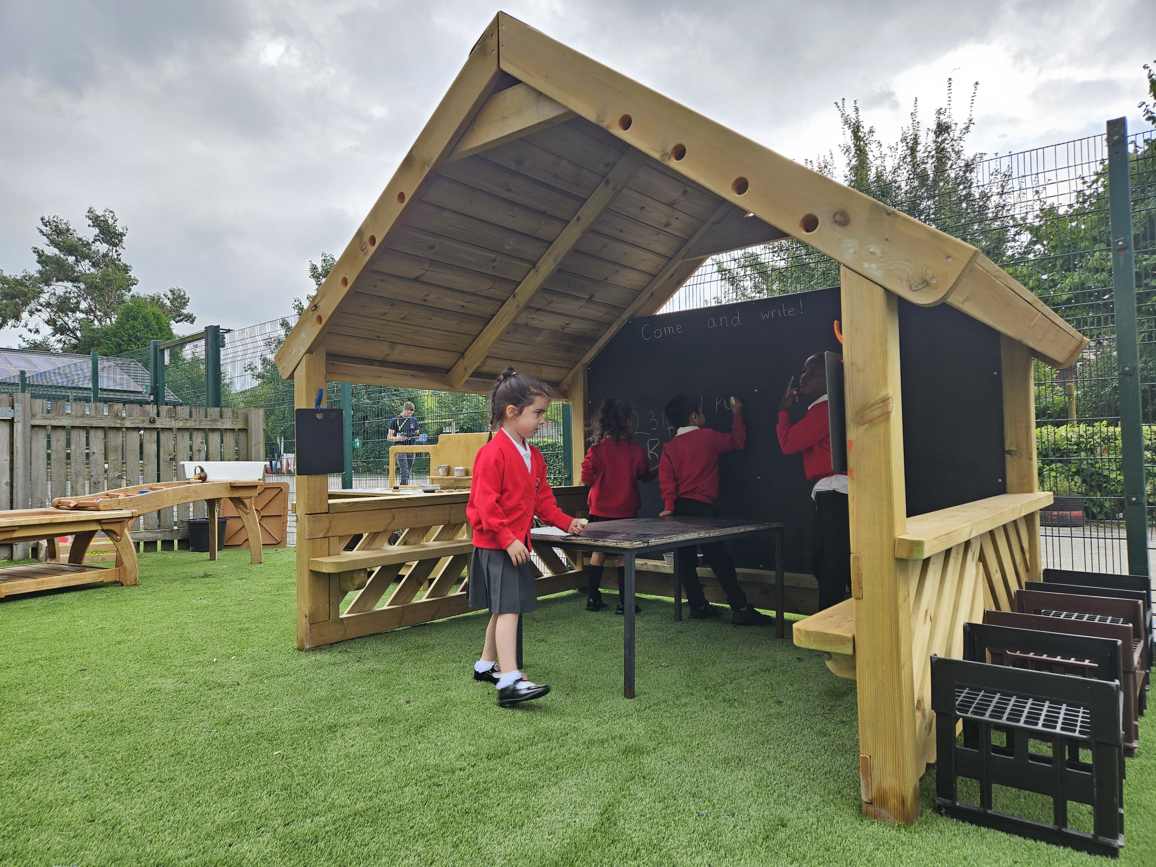4 children are underneath the Giant Playhouse and are writing on the big chalkboard in the background. The Giant Playhouse is on top of an artificial grass surface.