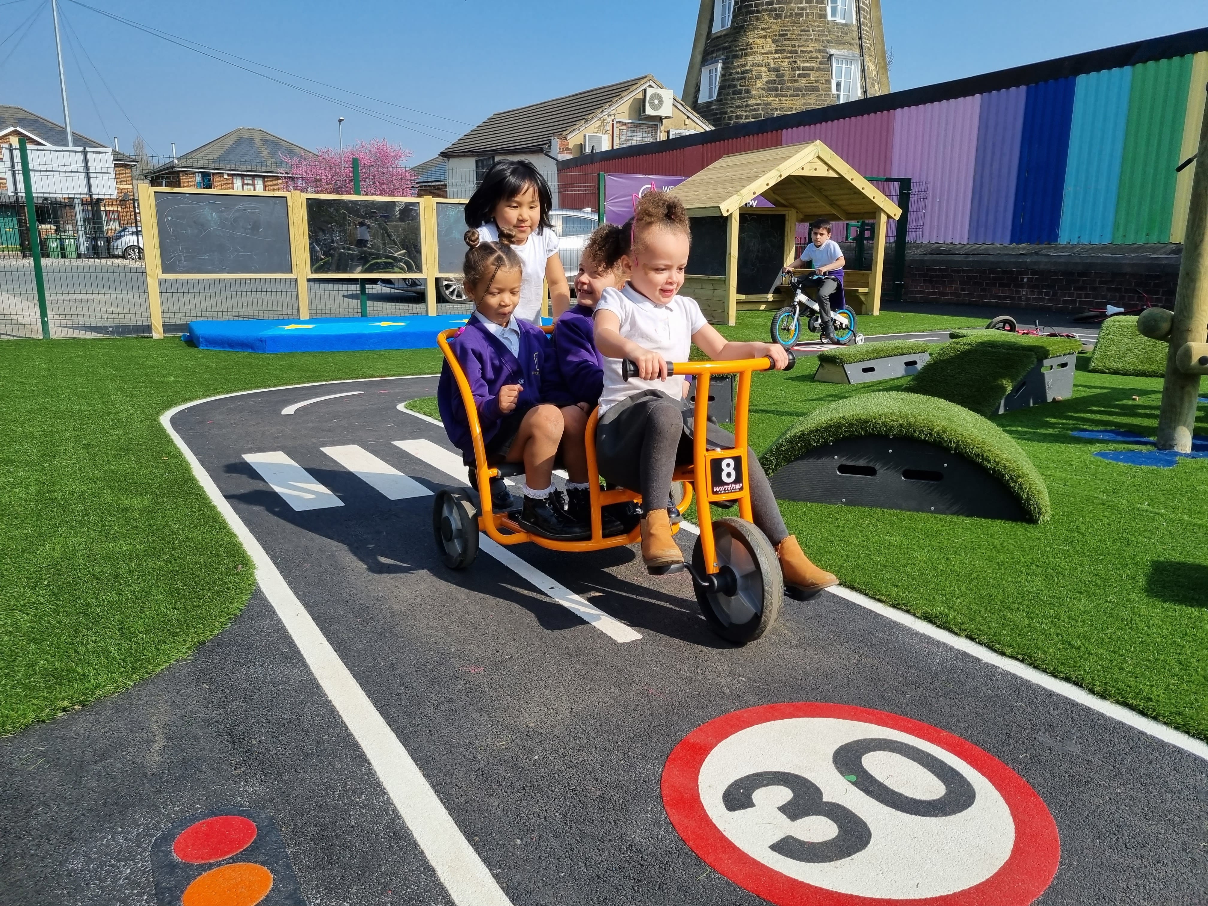 A child is riding a big bike, with 3 children on the back of it. They are riding on top of a road marking for a playground, with a child on another bike in the background.