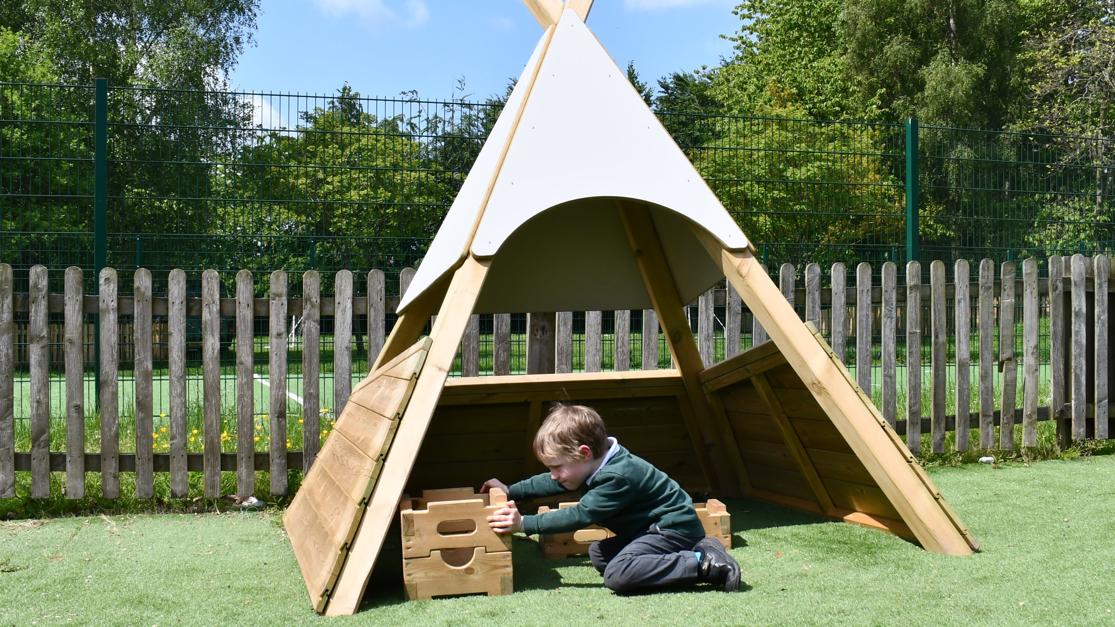 A child is sat under the Wigwam, playing with blocks and smiling. The Wigwam is surrounded by nature and the weather is nice.