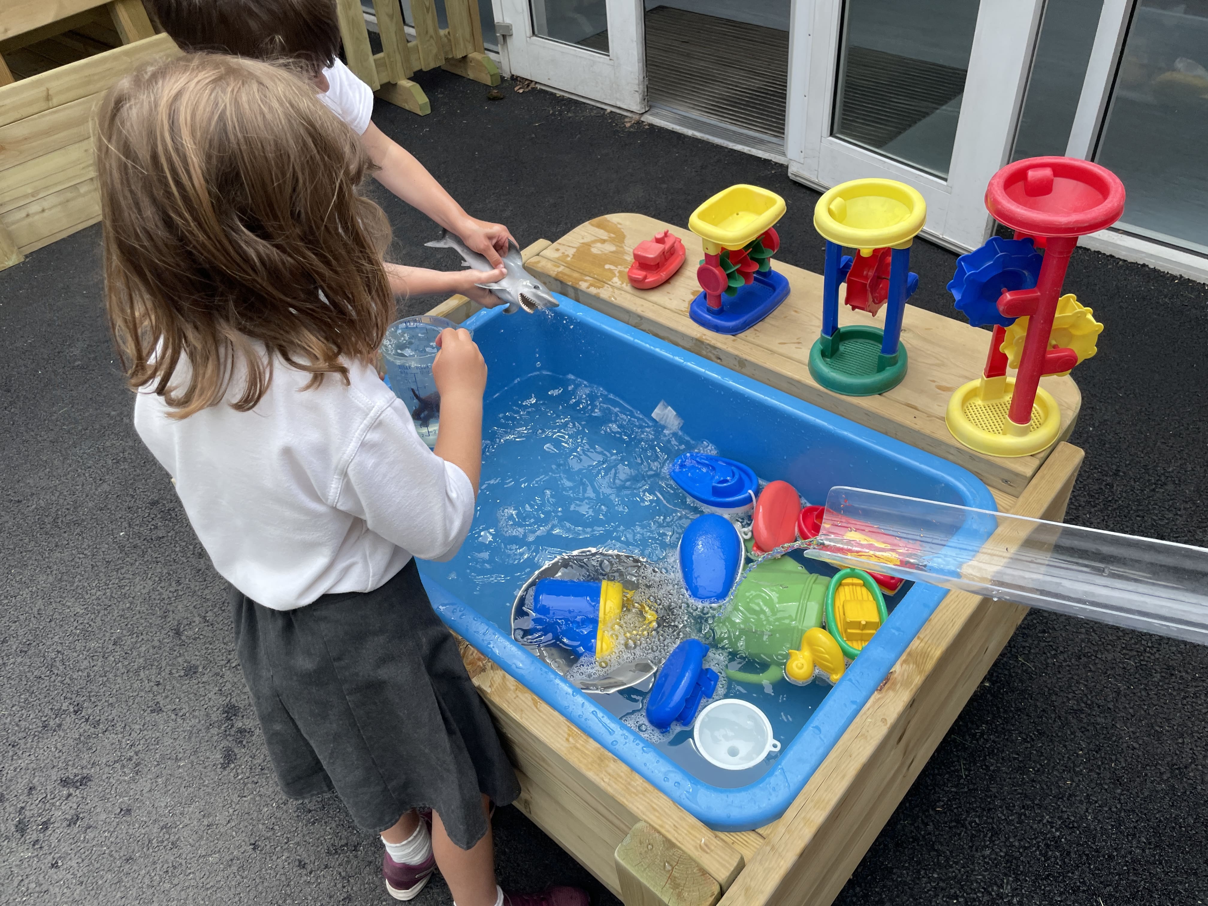 A girl and a boy are playing with a water table, which is a wooden box, with a plastic inside covering full of water. A wide variety of different water toys are placed around the area.