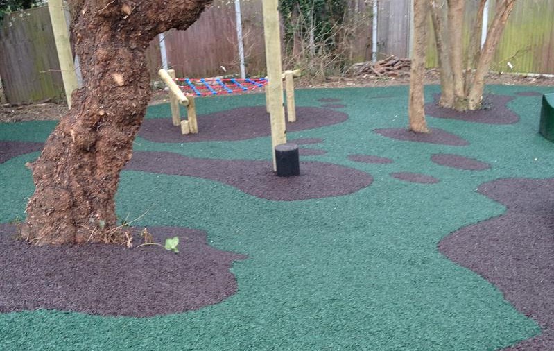 A picture showing a bonded rubber mulch play area. The colour of the mulch is in 2 different colours, green and brown. There's a tree to the left and climbing play equipment.