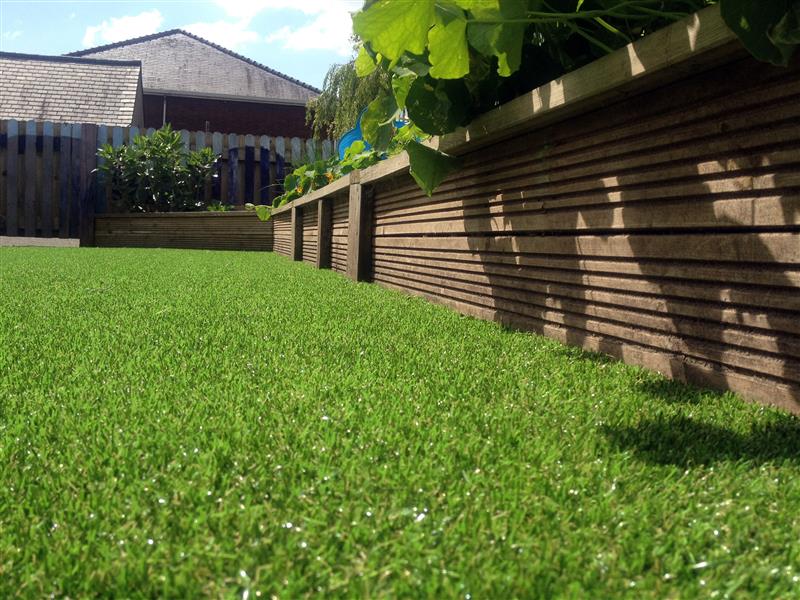 A close up photo of the Playturf, showing the quality of the artificial grass. Behind the grass are wooden planters, going around an area.