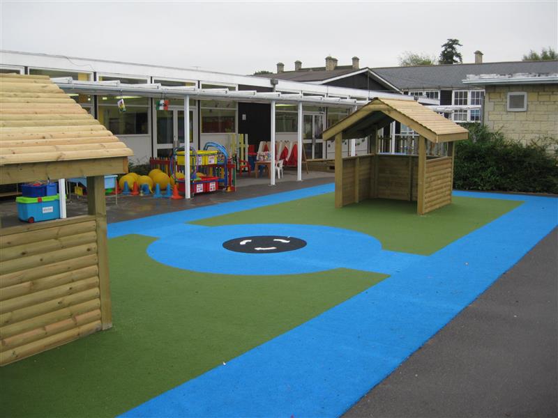 A saferturf playground with blue, green, black and white colours. The blue forms a rectangle with the green filling in the rectangle. A blue circle is in the centre of the green, with two blue rectangles connecting it to the big blue rectangle outline. A black circle is in the blue circle, with white arrows going around.
