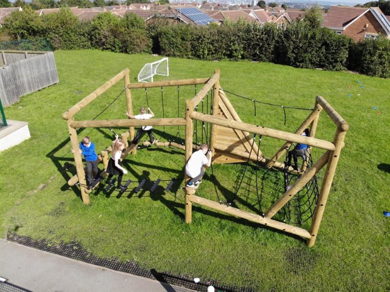 A climbing frame is placed on top of the safermats, which have grass growing through them. Grass is surrounding the equipment, with trees in the background (creating a natural feel).