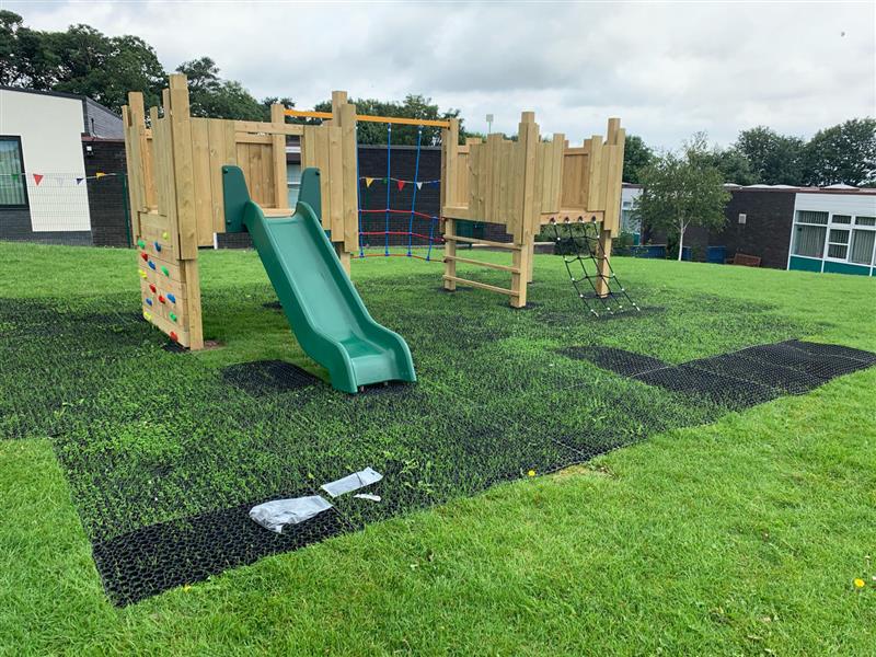 A play tower is on top of Safermats, with grass growing through them. The play tower is made of wood and has two parts that are connected by a climbing rope. The Safermats are visible and are black and grid like, with holes in them.