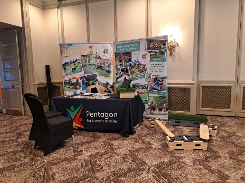 A picture showing our booth at the NMT Conference in 2023. A black clothes with the Pentagon Play logo is on top of a table, with two posters displayed behind it, showing the work we do. The table has other materials like brochures and pencils. Beside the table is a Play Builder set and Get Set, Go! Blocks