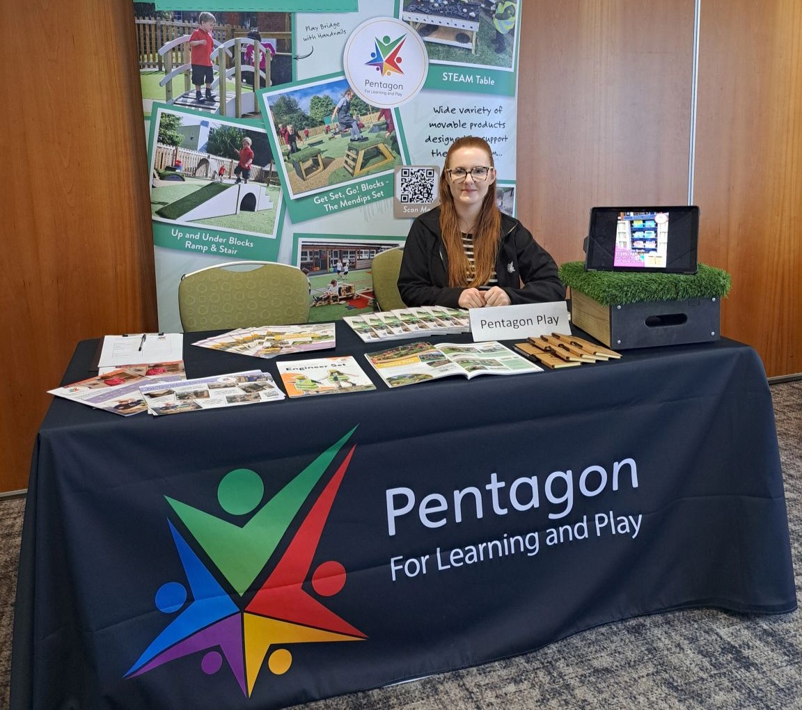 A table covered by a black cover with the Pentagon Play logo on the front. Behind the table is 2 seats, with a woman sat on the right. Behind the woman is a big poster showing all the services that Pentagon Play do. On the table is a bunch of different items like pencils, brochures, blocks and an iPad showing a presentation.