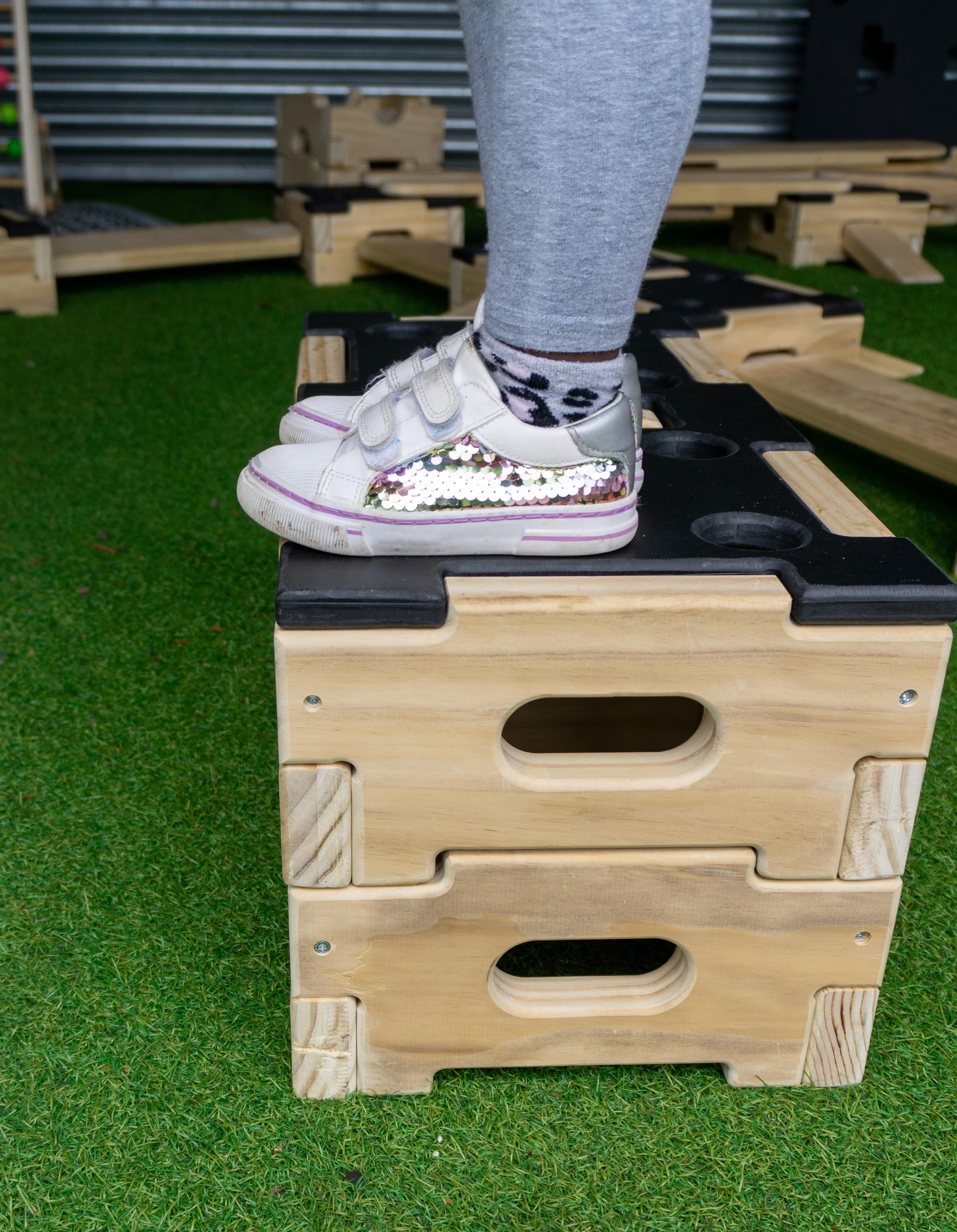 Accoya wood blocks are stacked on top of each other, which are a part of the play builder sets. A child is stood on top of both of the blocks, as the blocks are placed on top of artificial grass.,