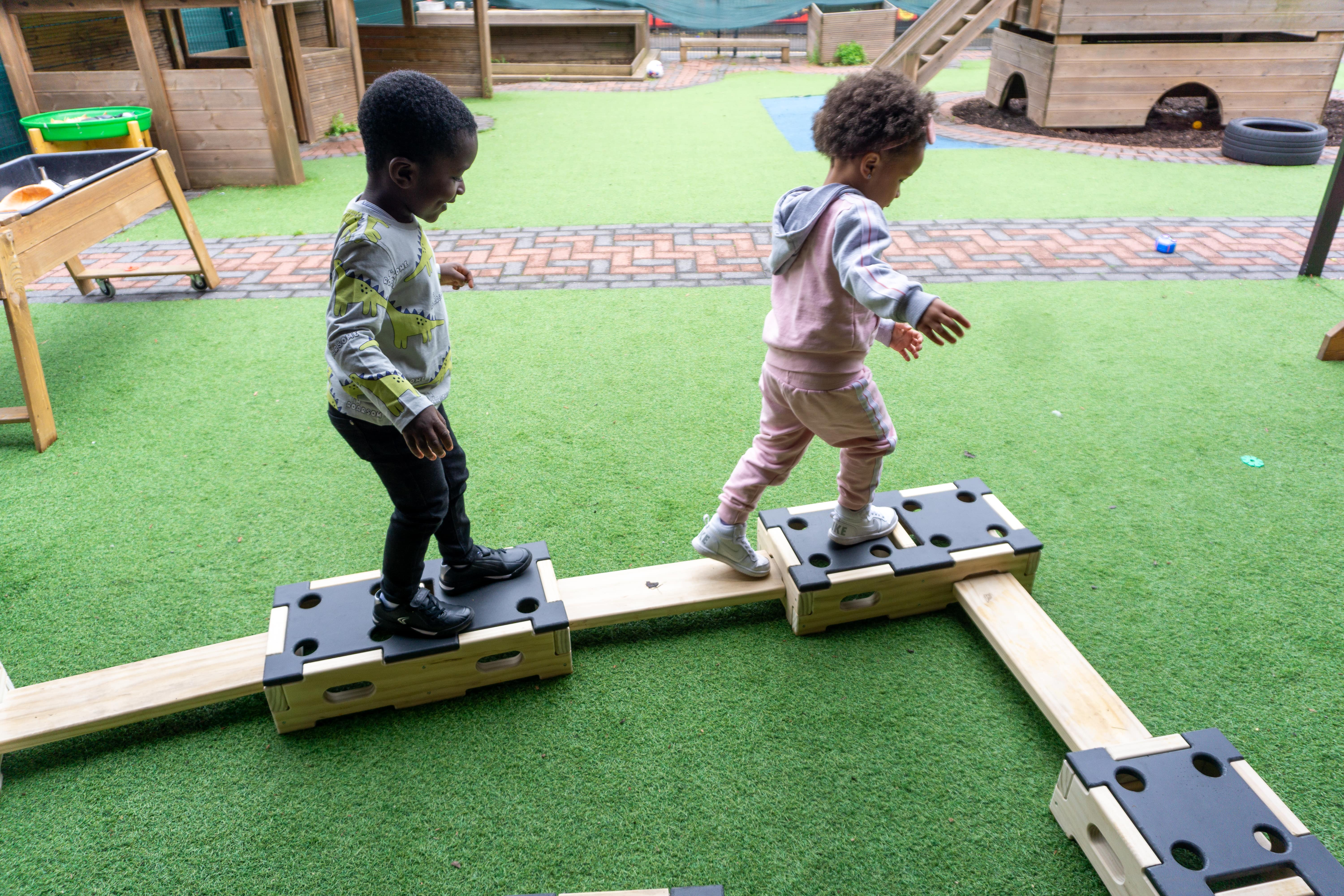 Two children are balancing across Accoya wooden beams. These beams are connected to Accoya wooden blocks which are all a part of the play builder sets. The obstacle course is laid on top of artificial grass